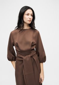 Γυναίκα με σκουρόχρωμα μαλλιά φορώντας καφέ φόρεμα με φουσκωτά μανίκια και πλατύ δετό ζωνάρι, κοιτάζει προς το πλάι μπροστά σε απλό φόντο.