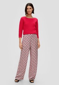 Roter gerippter Strickpullover mit einem Bootsausschnitt, kombiniert mit rosafarbigen weiten Hosen mit einem schwarzen Herzmuster, gestylt mit schwarzen Sandalen.