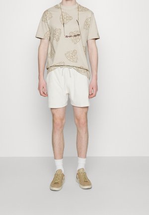 Man wearing beige tiger-print t-shirt, white drawstring shorts, white socks, beige sneakers, and sunglasses hanging on a cord around his neck.