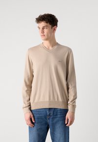 Beige V-kivágású pulóver hosszú ujjal, puha kötött anyagból készült, bordás részletekkel a mandzsettákon és az alján. Kék farmerral viselve.