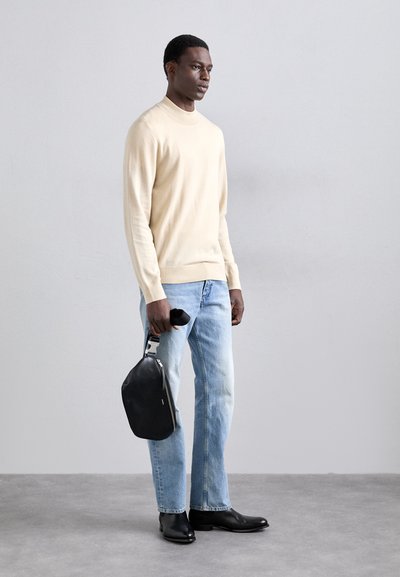 Beige Pullover, hellblaue Jeans und schwarze Lederstiefel. Eine schwarze Tasche mit einem silbernen Griff wird in der rechten Hand gehalten.