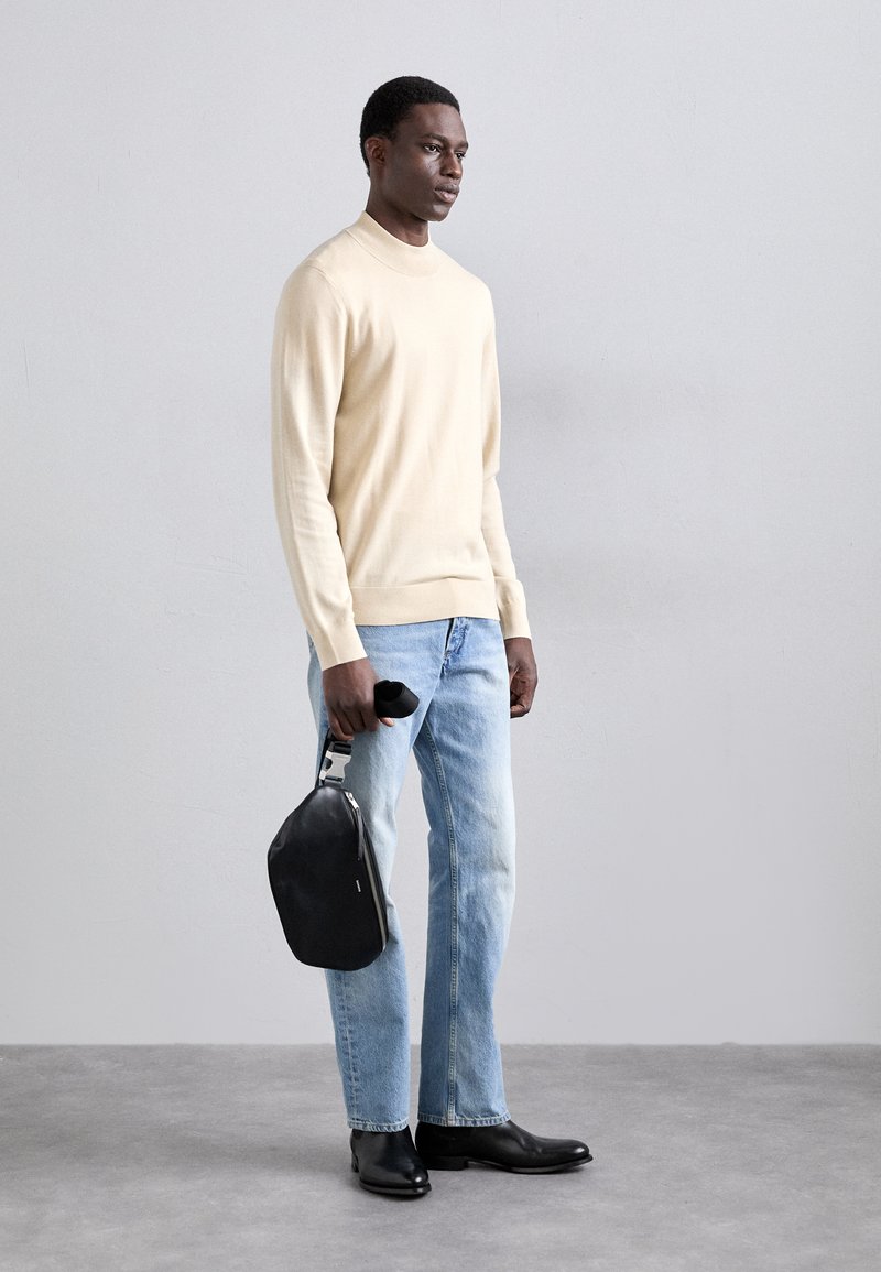 Un pull beige, un jean bleu clair et des bottes en cuir noires. Un sac noir avec une poignée en argent est tenu dans la main droite.
