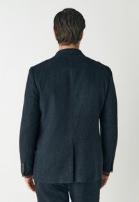 Tumedat sinised, kalasabamustrilised villased bleiser, millel on üks ventilatsioon, kitsad kraed ja nööbitavad mansetid, vaadatuna tagant.