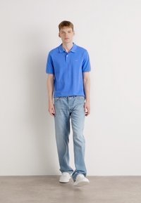 Blaues Baumwoll-Polohemd mit Kragen, kombiniert mit hellblauen Jeans und weißen Turnschuhen. Das Modell steht vor einem neutralen Hintergrund.