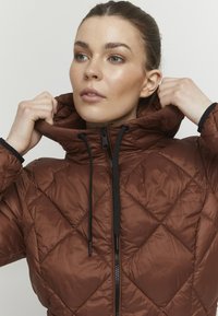 Veste matelassée marron avec un col haut, fermeture éclair noire et cordons de serrage. Présente un motif en losange et une texture lisse et brillante.