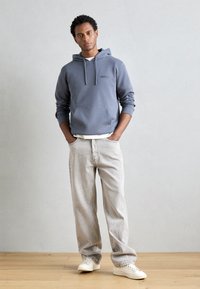 Siva kapuca z zadrgo, s sprednjim žepom, prekrita z belo majico. Svetlo obarvani, ravni kavbojki in bele superge.