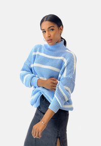 Helle blaue Strickjacke mit weißen horizontalen Streifen, die über einen hohen Kragen und lockere Ärmel verfügt, kombiniert mit einem dunklen Jeansrock.