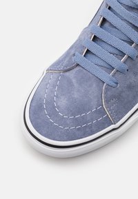 Blå mockaskor med rund tå, kontrasterande vit söm och platta snören. Har en svart gummisula. Inga synliga logotyper.