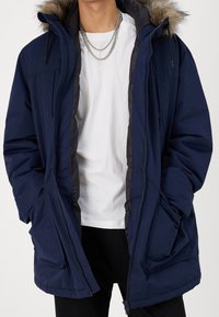 Mörkblå parka-jacka med pälsfodrad huva, dragkedja fram och flera fickor, med en slät textur och isolerad design.