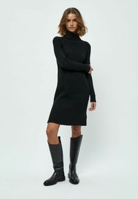 Abito in maglia a coste nera con collo alto e maniche lunghe, caratterizzato da una silhouette dritta e un orlo fino al ginocchio. Abbinato a stivali neri alti.