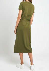 Femme debout, portant une robe midi à manches courtes vert olive et des baskets à plateforme blanches, de dos sur un fond uni.