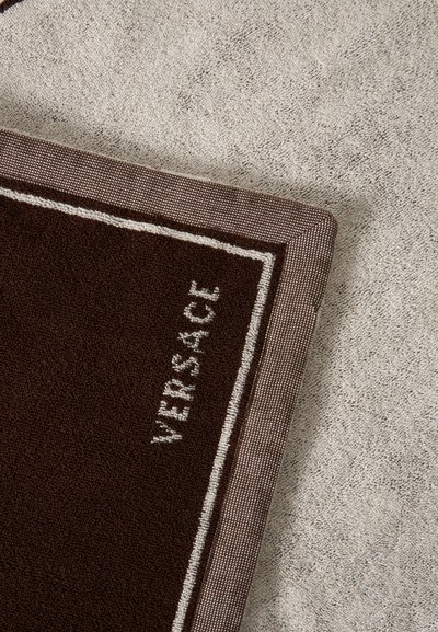 Roh hnedého obdĺžnikového uteráka s nápisom "VERSACE" a vzorovaným okrajom ležiaci na texturovanom béžovom koberci.