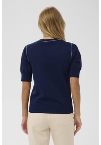 Moteris ilgais šviesiais plaukais, apsirengusi tamsiai mėlyna trumpomis rankovėmis megztiniu ir šviesiai bežė kelnėmis, atsukusi nugarą prieš vientisą foną.