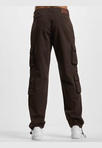 Bruine cargobroek van stof met vier zijzakken en een leren label op de achterste tailleband. Witte sneakers maken de look af.