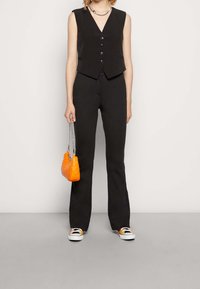Svart väst utan ärmar med V-halsringning och fem knappar, tillsammans med svarta utsvängda byxor. Accessoarer inkluderar en liten orange vadderad handväska.