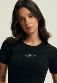 Czarny, prążkowany t-shirt z okrągłym dekoltem i wyśrodkowanym białym tekstem: "NAPRAWDĘ WYGLĄDA DOBRZE NA TOBIE, POZOSTAŃ SPOKOJNY, BLOODVII". Krótkie rękawy.