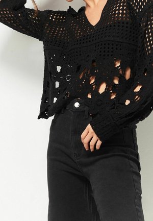 Femme portant un haut à manches longues en crochet noir avec des motifs floraux et un jean noir taille haute, posant avec une main près de la taille.