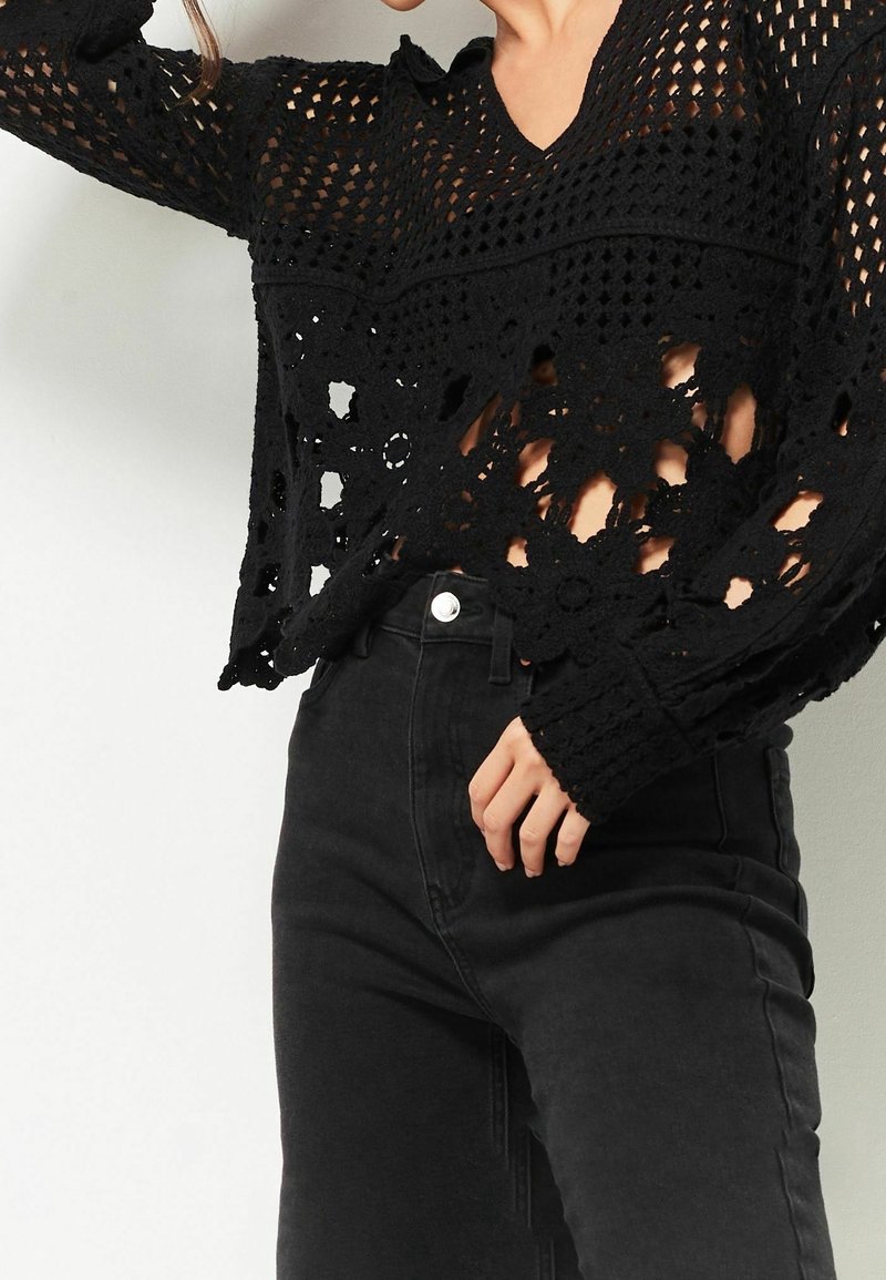 Femme portant un haut à manches longues en crochet noir avec des motifs floraux et un jean noir taille haute, posant avec une main près de la taille.