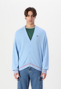 Cardigan azzurro chiaro con scollo a V, chiusura a bottoni e polsini a righe rosse. Texture a coste e un piccolo logo verde sul petto. Indossato sopra una camicia verde.
