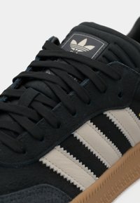 Black suede sneaker with three beige stripes, black laces, and a textured gum sole. Features a branded tongue label.