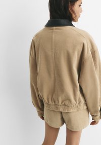 Beige corduroy takki mustalla nahka-kauluksella, jossa on rento istuvuus ja joustava helma. Mallilla on päällään yhteensopivat shortsit.