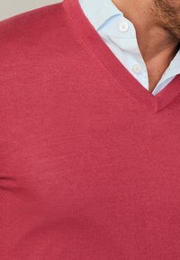 Roter Strickpullover mit V-Ausschnitt, der eine glatte Textur und eine enge Passform aufweist. Über einem hellblauen Button-Up-Hemd getragen.