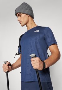 Sötétkék teljesítmény t-shirt, texturált mintával és fehér logóval, szürke kötött sapkával párosítva; trekking botokat tart a kezében.
