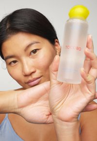 Bouteille en plastique transparent avec un bouchon jaune, étiquetée "TOCOBO" en rouge et bleu, tenue par une personne portant un haut bleu clair, montrant une texture légère et aqueuse.