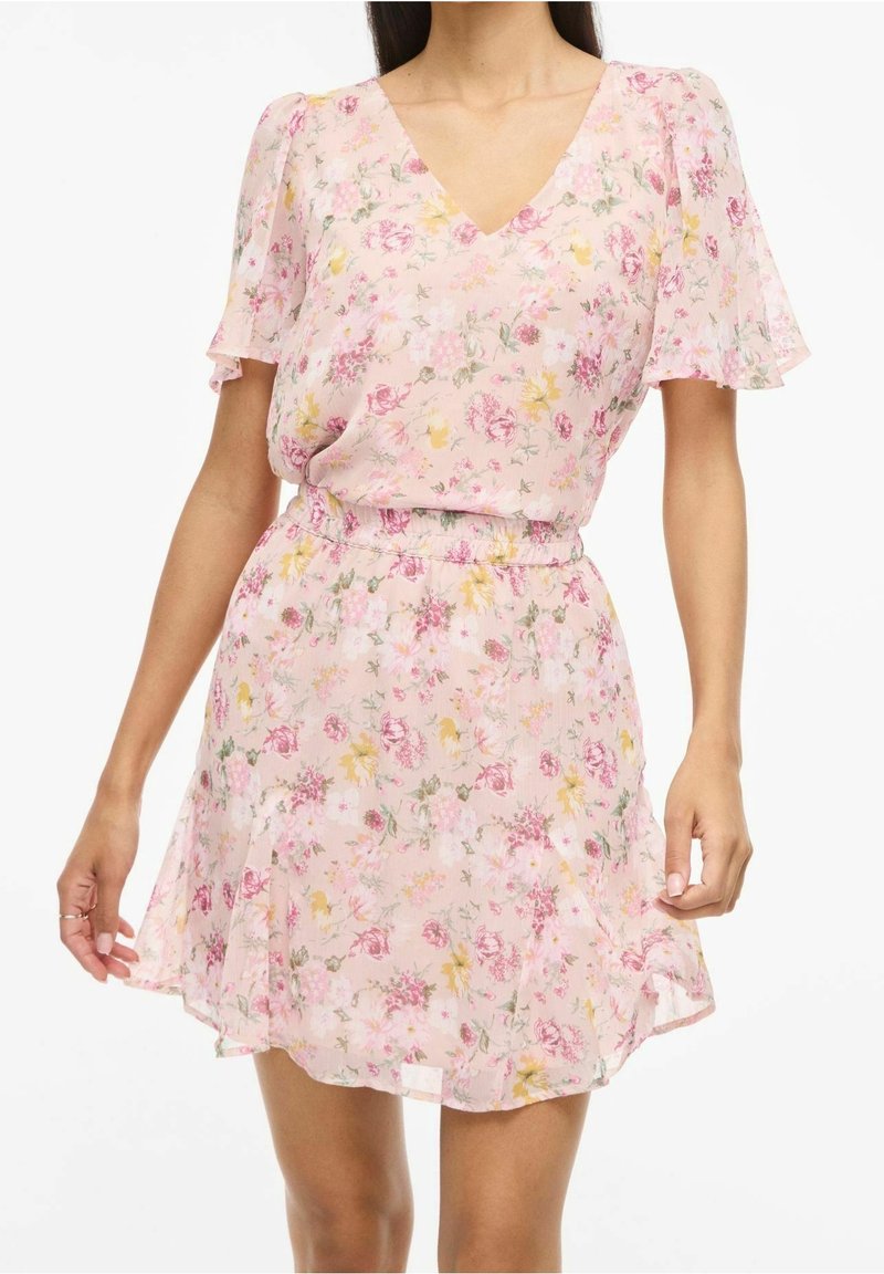 Femme portant une robe courte rose à fleurs avec manches courtes, un décolleté en V et une taille cintrée, debout devant un fond blanc.