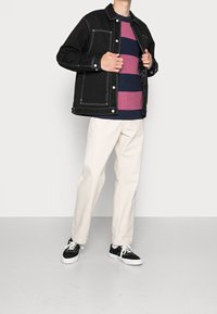 Black denim jacket with white stitching, striped burgundy and navy sweater, cream pants, and black sneakers, displayed against a neutral background.
