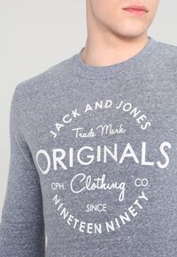 Uomo che indossa un felpa grigia con testo bianco che recita "Jack and Jones Originals Clothing Co. dal Millenovecentonovanta"