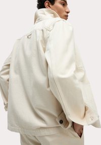 Cream-colored denim jacket with a collar, large sleeves, and buttoned cuffs. Features double-stitch detailing and minimalistic design.