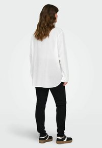 Camicia bianca a maniche lunghe con orlo curvo, abbinata a pantaloni neri slim-fit e sneakers nere con dettagli bianchi. Design semplice e casual.