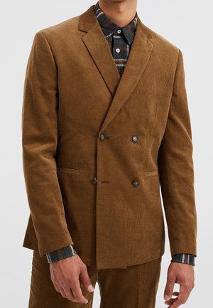 Man draagt een bruin corduroy dubbelbreasted colbert over een donker geruit overhemd, staand tegen een effen achtergrond.