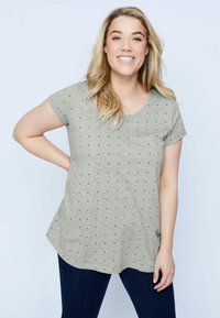 Camiseta de manga corta de color gris claro con cuello redondeado y patrón de lunares negros. Tejido suave con un corte relajado, combinada con jeans de color azul oscuro.