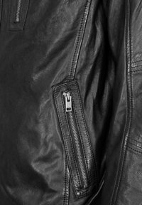 Svart läderjacka med en synlig dragkedjeficka och strukturerad yta. Utmärkande sömmar längs sömmarna och släta dragkedjekomponenter.