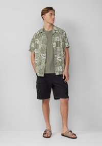 Korte mouwen, groene gestreepte shirt over een effen olijfgroene t-shirt, gecombineerd met zwarte cargo shorts en bruine slipper sandalen.