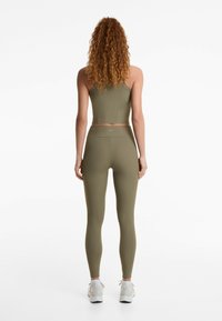 Top scurt cu dungi de culoare verde măslinie și leggings, design ajustat, material texturat, asortat cu adidași albi. Detaliu minimal de marcă vizibil.