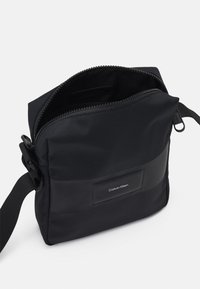 Borsa a tracolla nera con tessuto testurizzato, chiusura a zip e un'etichetta logo in rilievo su un pannello liscio. Inclusa una tracolla regolabile.