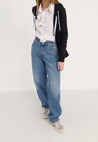 Jeans de tiro alto en azul claro con un ajuste holgado, combinados con una camisa blanca de botones atada a la cintura y una sudadera con cremallera negra.