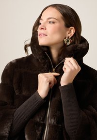 Brązowy płaszcz z sztucznego futra z wysokim kołnierzem, fakturowanym materiałem i zapięciem na zamek. Model nosi sweter z prążkowanymi rękawami oraz ozdobną kolczyk.