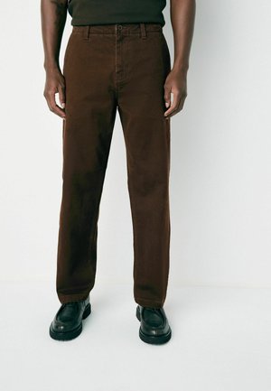 Man wearing straight-fit brown pants and black leather shoes standing against a plain white background.