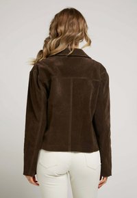 Femme avec des cheveux châtain clair et ondulés porte une veste en daim marron foncé et un pantalon blanc ajusté, se tenant devant un fond clair et uni.