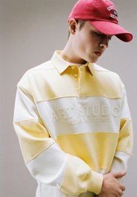 Jeune personne portant un sweat-shirt rayé jaune et blanc avec le texte "ART STUDIO Creative Department" et une casquette de baseball rouge, regardant vers le bas.