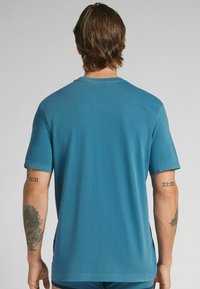 Modré tričko s krátkým rukávem z měkkého materiálu, s kulatým výstřihem a bočními rozparky. Hladká textura s tetováním viditelným na levém rameni.
