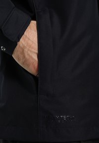Crna Helly Tech jakna s teksturiranim materijalom, skrivenim džepom i ukrasnim logom u sjajnom finišu na rubu.