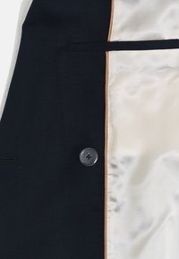 Navy blazer met een satijnen crèmekleurige voering, voorzien van een zwarte knop, een borstzak en een contrasterende tan afwerking langs de rand.