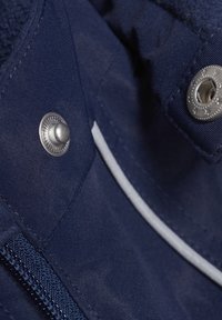Marinblå tyg med en mjuk textur, prydd med silverfärgad metallknapp och dragkedjedetaljer. Inkluderar en kontrasterande vit accent.