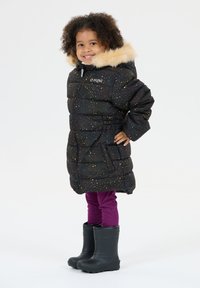 Lille barn smilende, iført en sort puffjakke med multicolor prikker, beige pelshætte, lilla leggings og sorte gummistøvler.