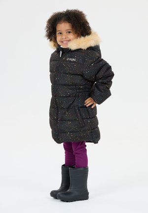 Lille barn smilende, iført en sort puffjakke med multicolor prikker, beige pelshætte, lilla leggings og sorte gummistøvler.